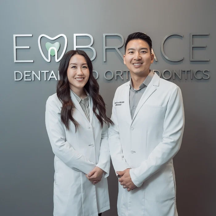 Dr. Christina S Lee and Dr. Peter Lee in white coats standing in front of a Embrace Dental and Orthodontics sign
